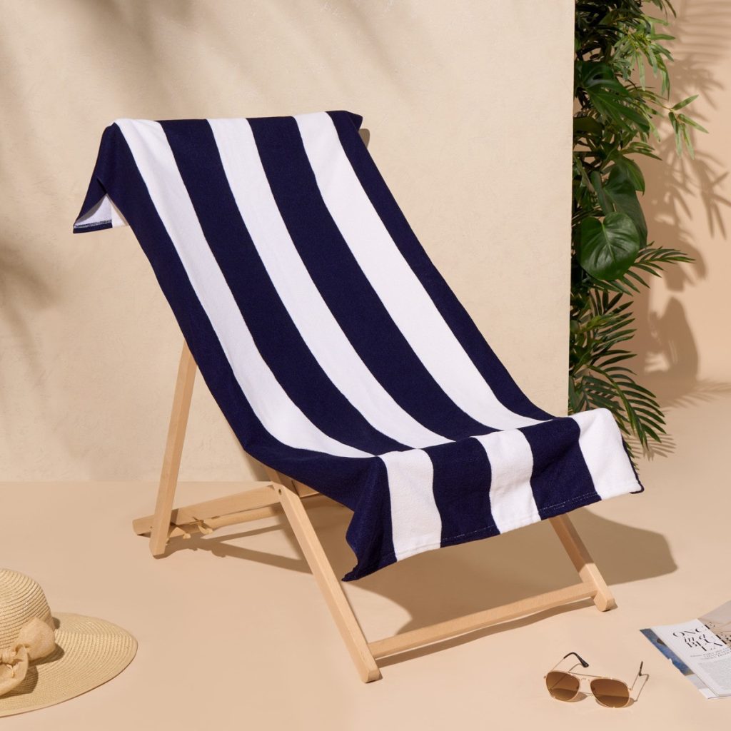 A vertical striped beach towel is displayed in the image, featuring a blue stripe and a white stripe. The towel is folded and placed on a surface, with a hat nearby. The background is composed of a wall.