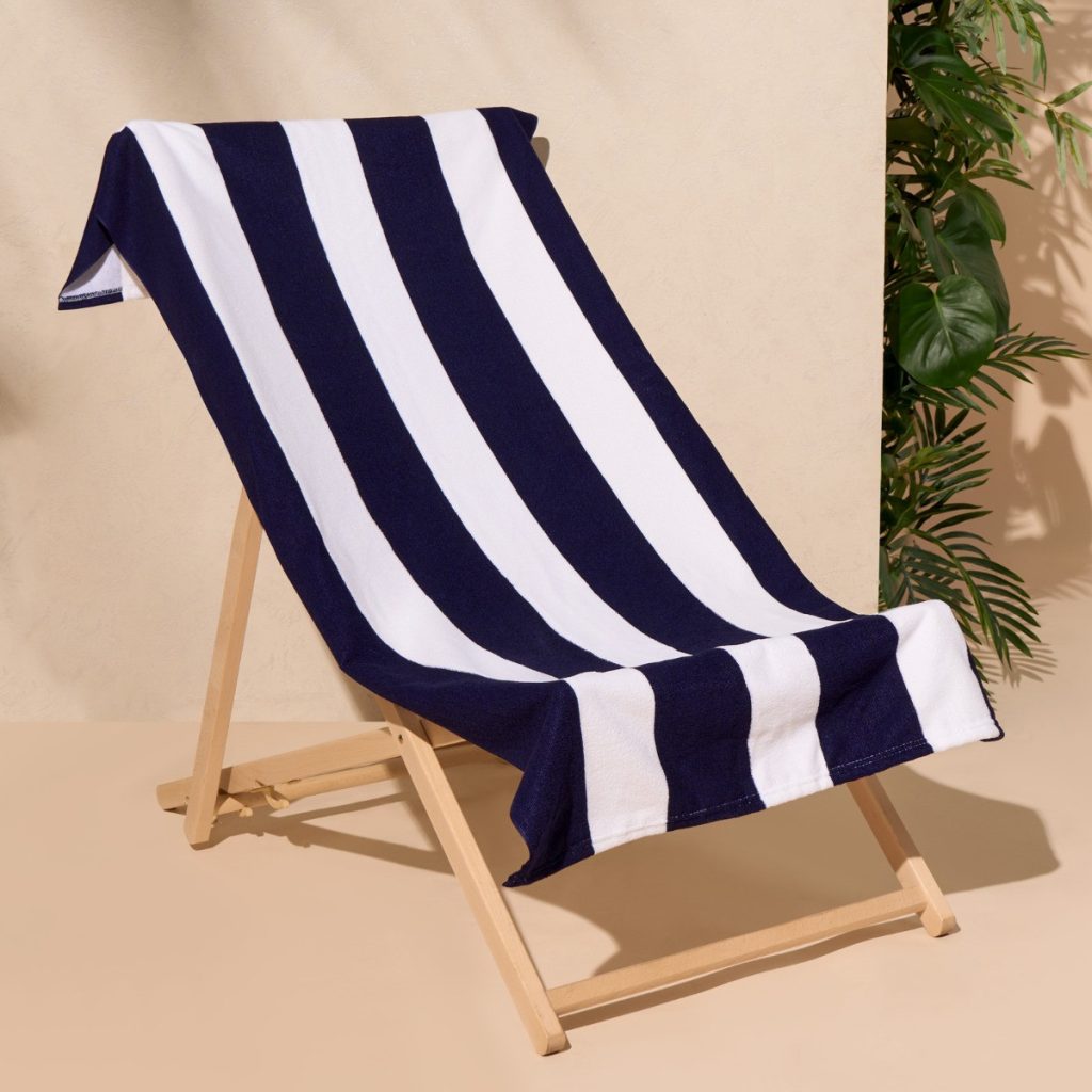 A blue and white striped towel is depicted in this image, with a wooden stand in the foreground. The background features a cream-colored wall and a plant.