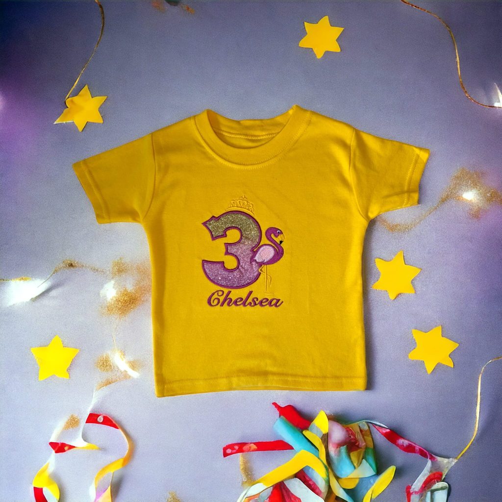 A yellow t-shirt is displayed in the image, featuring the number three and a pink flamingo design. The shirt is placed on a purple surface, with a few yellow stars visible in the background.