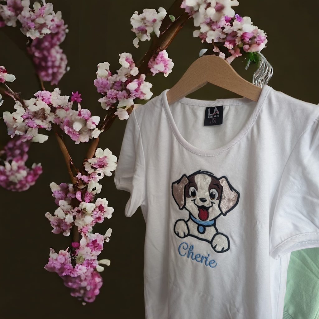 A white t-shirt is depicted in this image, featuring a cartoon image of a dog and the name "cherie. " the shirt is hanging on a hanger, and a pink flower is visible in the background.