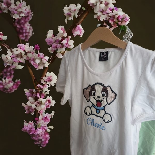 A white t-shirt is depicted in this image, featuring a cartoon image of a dog and the name "Cherie." The shirt is hanging on a hanger, and a pink flower is visible in the background.
