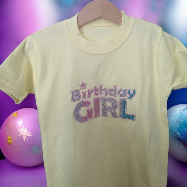 A t-shirt is displayed in the image, featuring text and balloons. The balloons are blue and pink in color. The background is a mix of purple and pink.