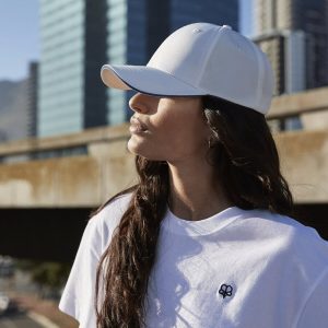 A woman is standing in the image, dressed in a white T-shirt and a cap. The background features a bridge, a building, and the sky.