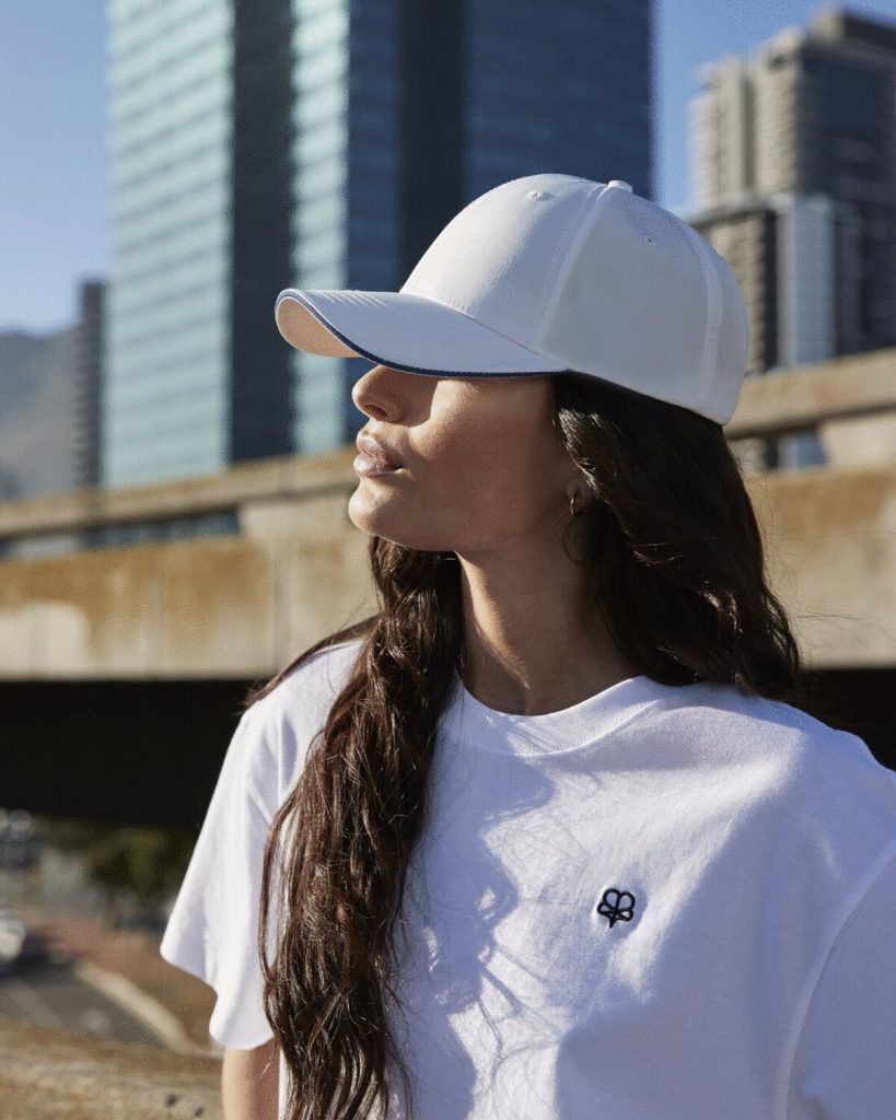 A woman is standing in the image, dressed in a white t-shirt and a cap. The background features a bridge, a building, and the sky.