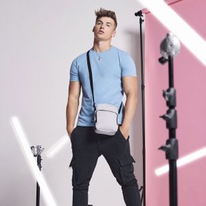 A man is standing in the image, dressed in a blue t-shirt and black pants. He is carrying a bag in his hand. The background features a pink wall and several black poles.