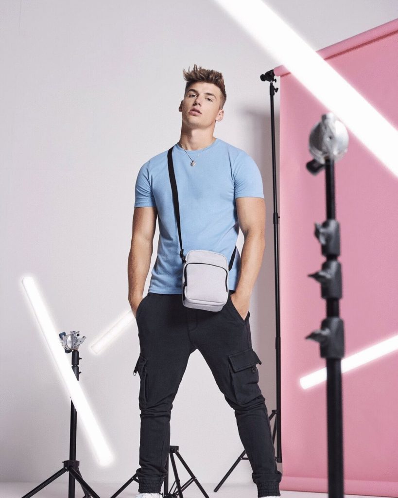 A man is standing in the image, dressed in a blue t-shirt and black pants. He is carrying a bag in his hand. The background features a pink wall and several black poles.