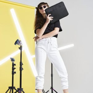 A woman is standing in the image, holding a black laptop bag. The background features a yellow wall with a few lights, and a white wall with a yellow background.