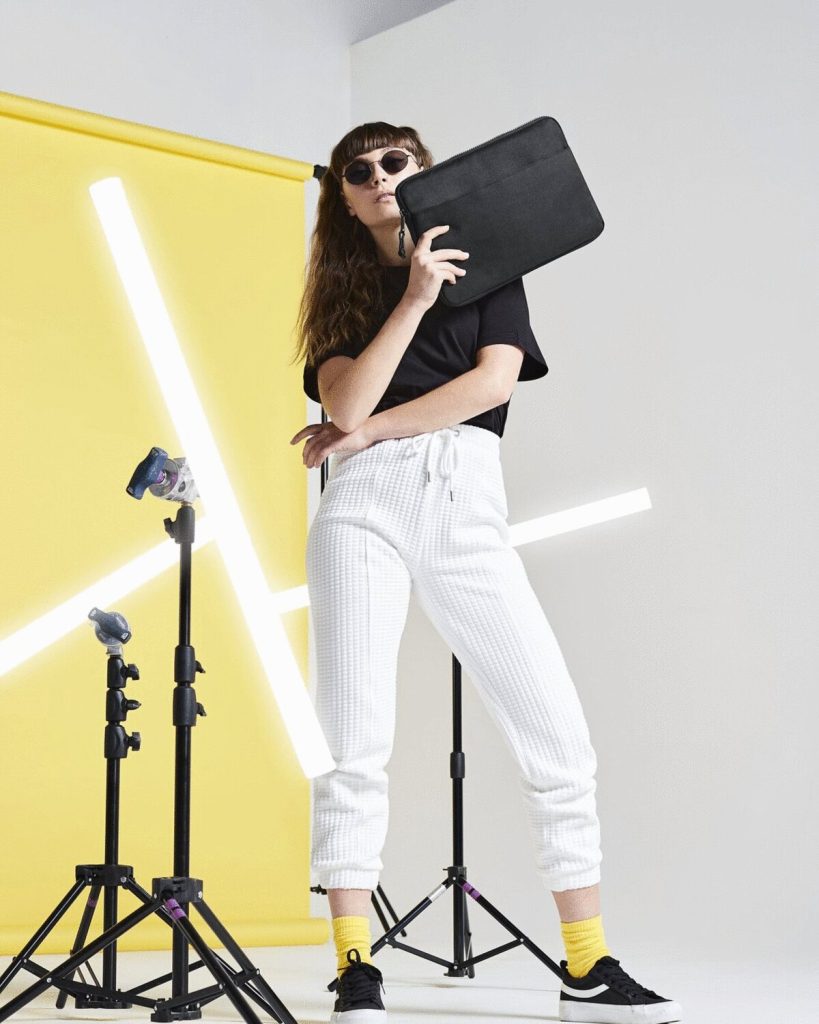 A woman is standing in the image, holding a black laptop bag. The background features a yellow wall with a few lights, and a white wall with a yellow background.