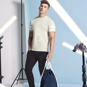 A man is standing in the image, dressed in a white t-shirt, black pants, and a blue bag. The background features a white wall and a few black objects.