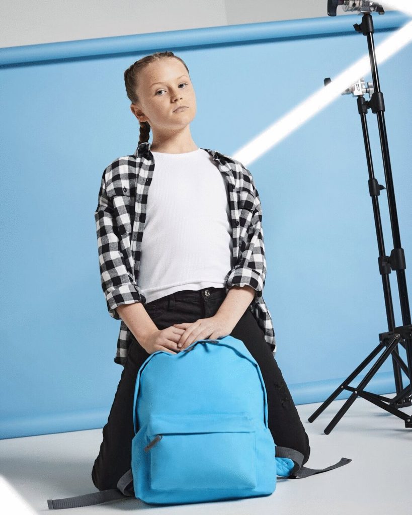 A girl is seated on the floor, wearing a white t-shirt and a black jacket. She is holding a blue bag in her hands. The background is a solid blue color. The image is taken from a close-up perspective.