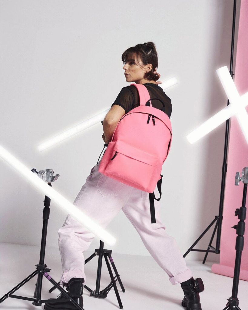 A woman is standing in the image, dressed in a black t-shirt and pink pants. She is holding a backpack in her hand. The background features a white wall and a pink light.