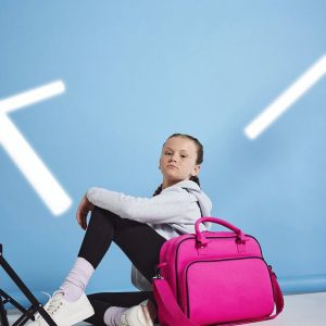 A girl is seated on the floor, holding a pink bag in her hands. The background features a blue wall, and a black stand is situated to the right side of the image.