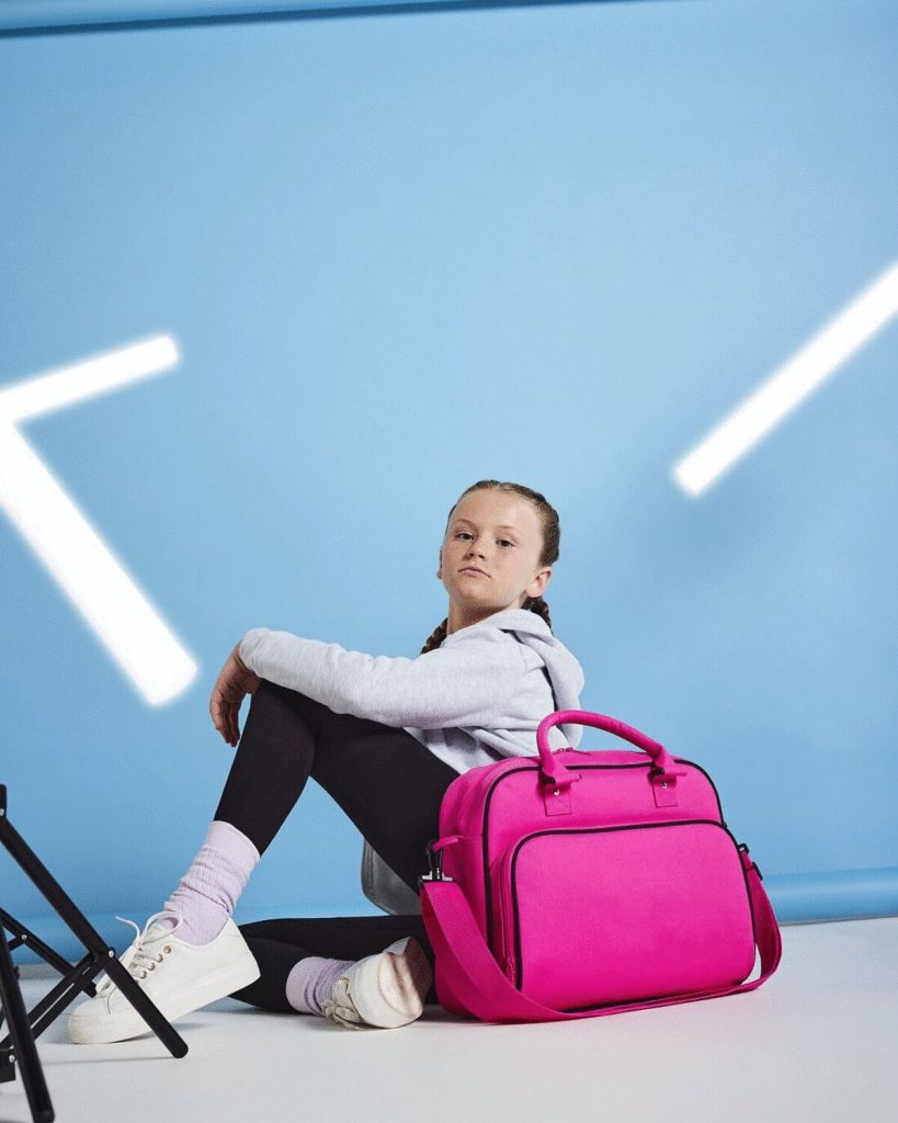 A girl is seated on the floor, holding a pink bag in her hands. The background features a blue wall, and a black stand is situated to the right side of the image.