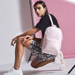 A woman is depicted in this image, wearing a black t-shirt and holding a pink backpack. The background features a pink wall, and the image is slightly blurry in the background.