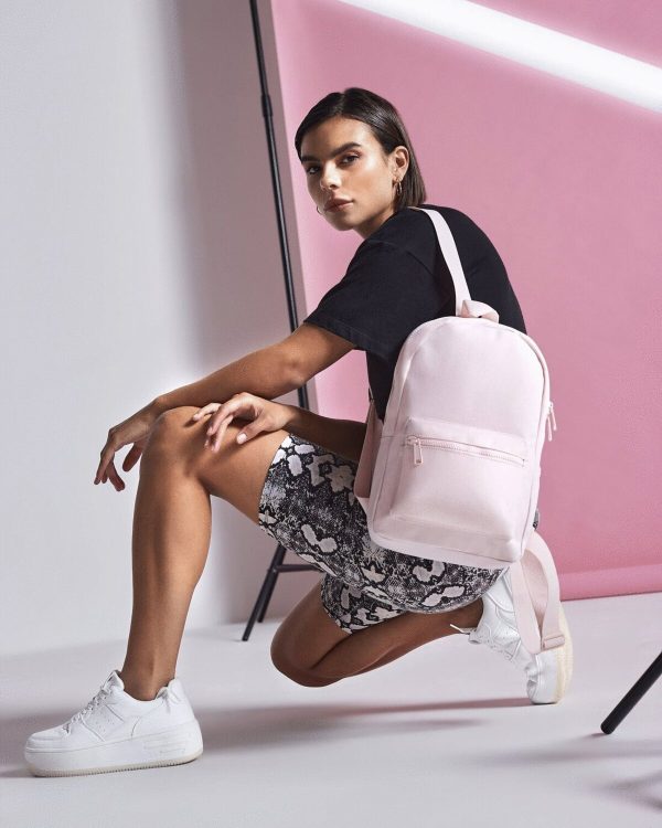 A woman is depicted in this image, wearing a black t-shirt and holding a pink backpack. The background features a pink wall, and the image is slightly blurry in the background.