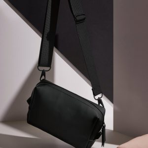 A black bag is the central focus of this image, with a pink background. The bag is placed on a white surface, and a pink object is situated to the right of the bag.