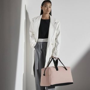 A woman is standing in the image, dressed in a black top and a white coat. She is holding a pink bag in her hand. The background is white, and the image appears to have been taken in a dimly lit environment.