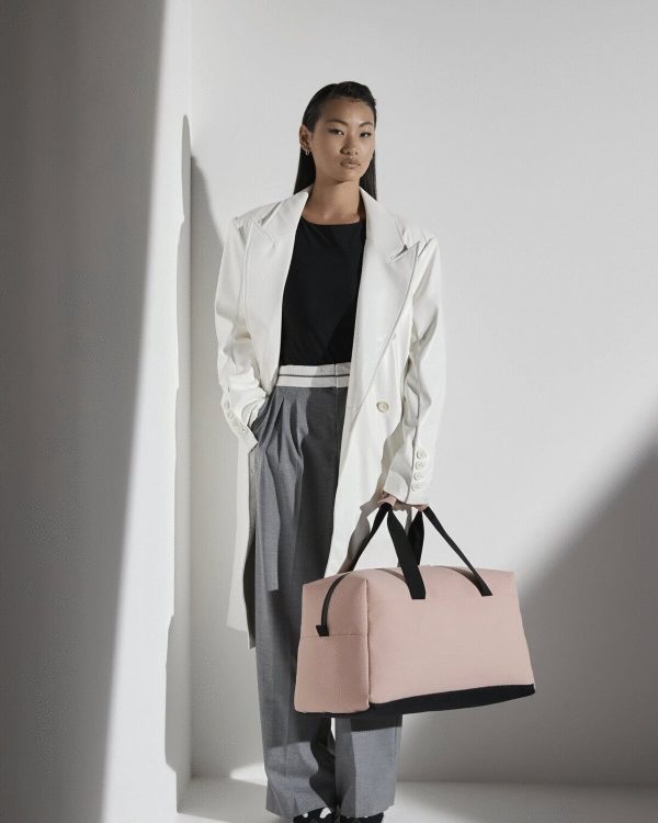 A woman is standing in the image, dressed in a black top and a white coat. She is holding a pink bag in her hand. The background is white, and the image appears to have been taken in a dimly lit environment.