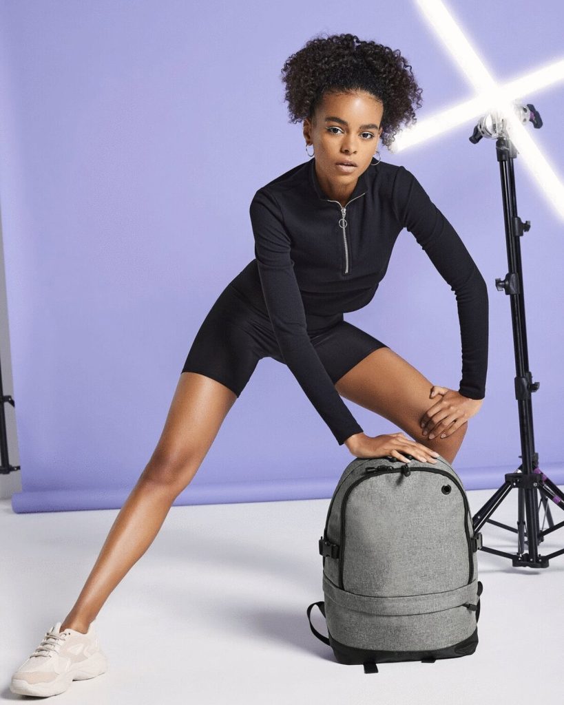 A woman is standing in the image, dressed in black. She is holding a grey backpack in her hands. The background features a white wall, and the image appears to have been taken in a studio setting.