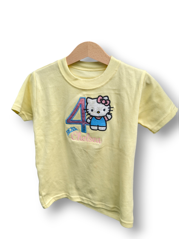 A child's t-shirt is depicted in this image, featuring a Hello Kitty design and the number four. The shirt is hanging on a hanger, and the background is white.