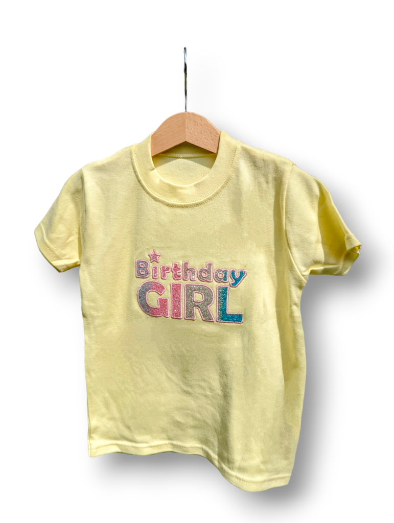 A yellow t-shirt is displayed in the image, featuring the text "birthday girl. " the shirt is adorned with sparkly letters and appears to be hanging on a wooden hanger.