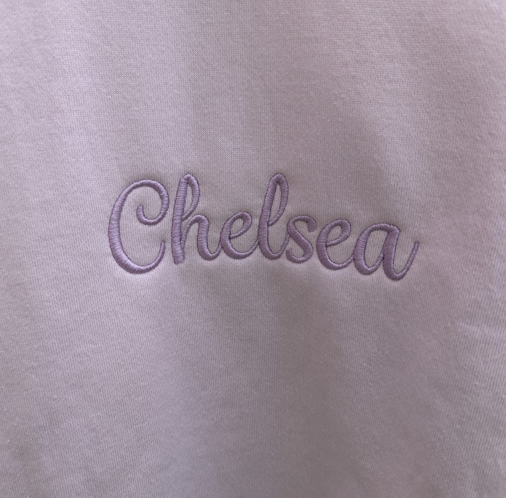 A white t-shirt is the central focus of this image, featuring the name "chelsea" written in purple. The background is white, and the image is taken from a close-up perspective.