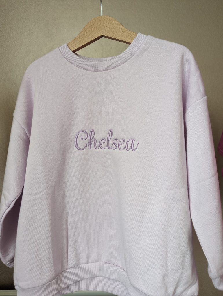 A purple sweatshirt is depicted in this image, featuring the name "chelsea" embroidered on it. The sweatshirt is hanging on a wooden hanger, and a white wall is visible in the background.