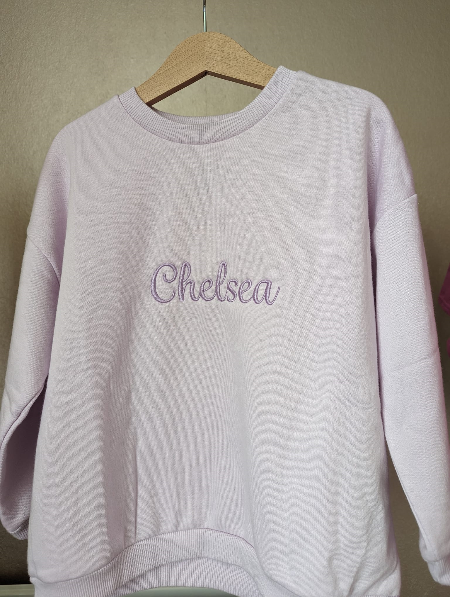 A purple sweatshirt is depicted in this image, featuring the name "Chelsea" embroidered on it. The sweatshirt is hanging on a wooden hanger, and a white wall is visible in the background.