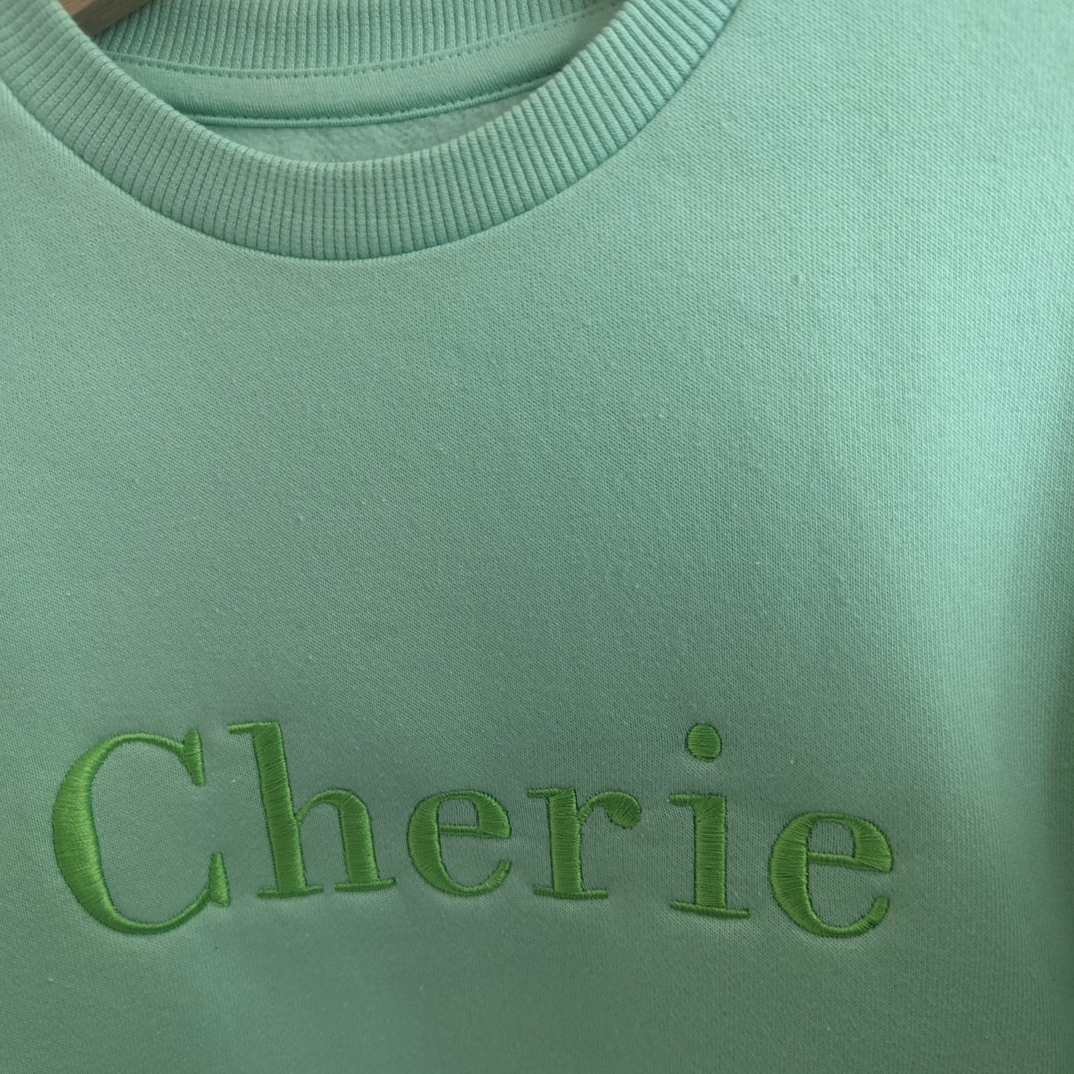A close-up image of a sweatshirt is presented, featuring the name "Cherie" embroidered on it. The sweatshirt is in a light green color, and the name is written in white letters.