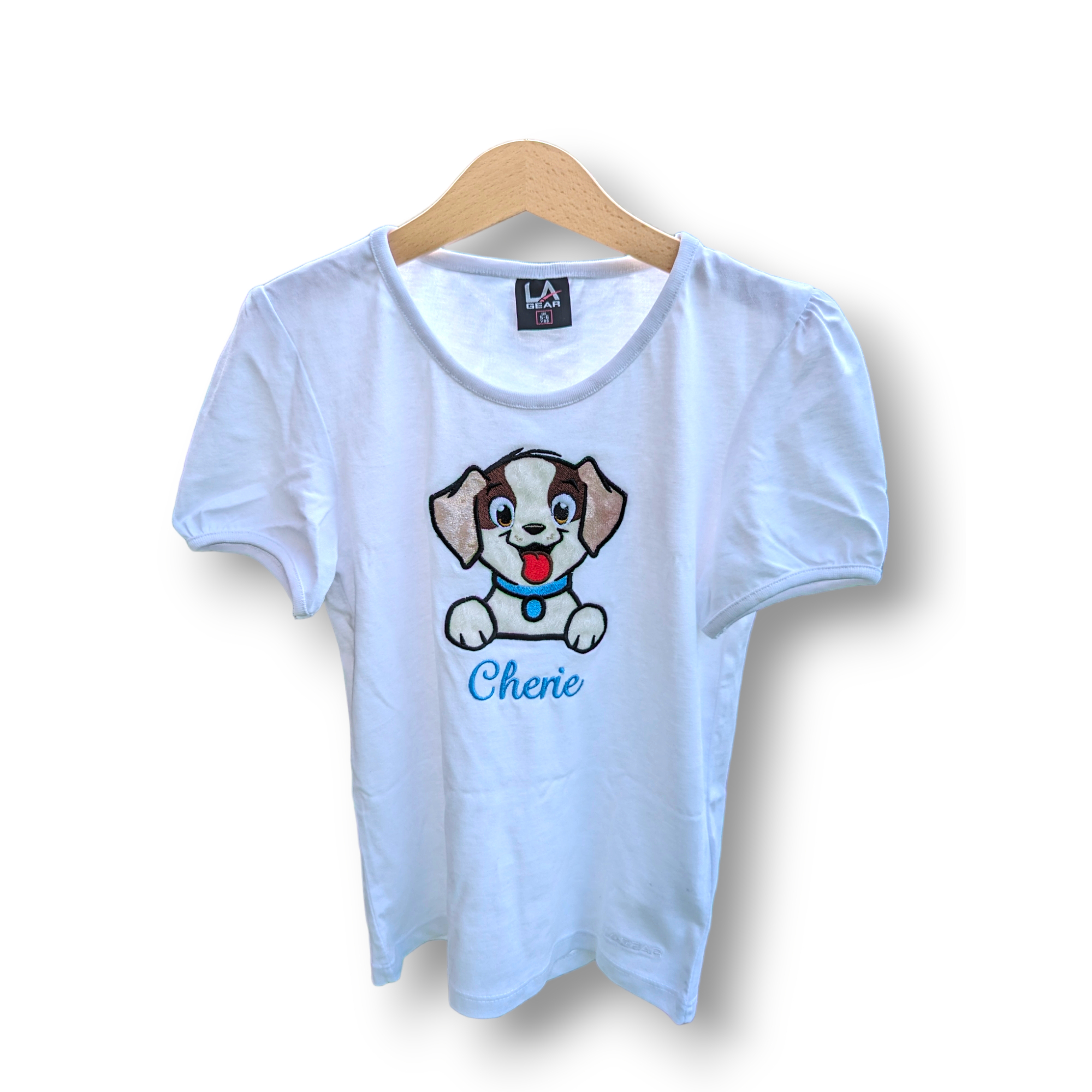 A white t-shirt is depicted in this image, featuring a cartoon image of a dog and the name "Cherio." The shirt is hanging on a wooden hanger.