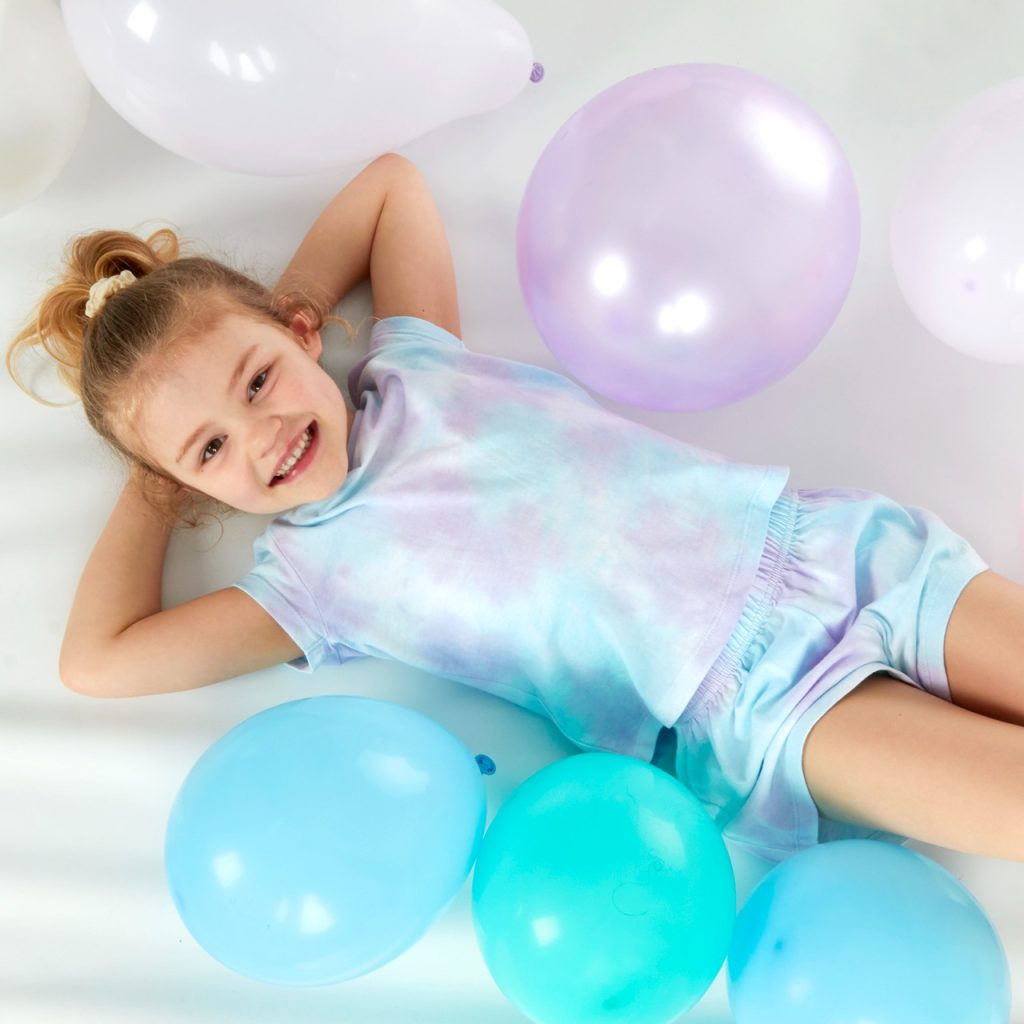 A girl is lying on a white surface, surrounded by balloons in various colors, including blue, pink, and purple. The background is white.