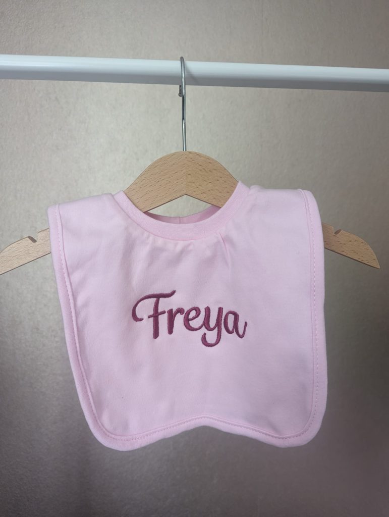 A pink-colored bib is the central focus of this image, with the word "freya" written on it. The bib is hanging on a white rod, which is attached to a white wall.