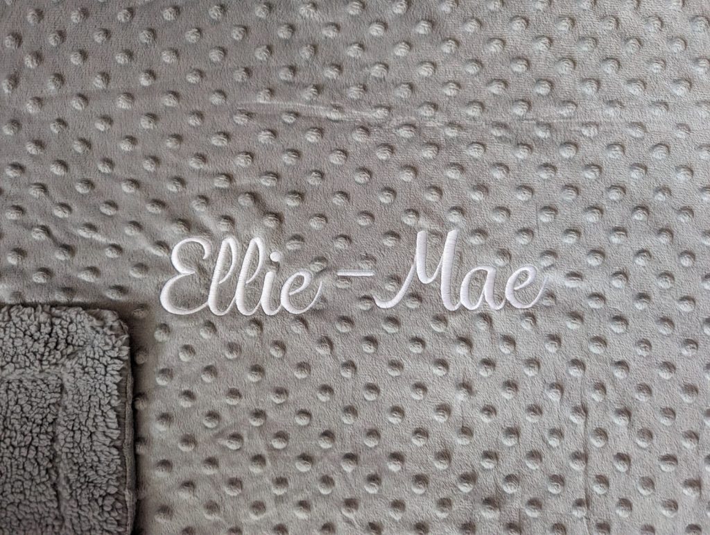 A grey-colored blanket is depicted in this image, featuring a white border and text. The blanket appears to be embroidered, with a few white dots visible on the surface.