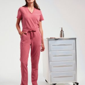 A woman is standing in the image, dressed in a pink top and pants. A white cabinet is situated behind her, and a bottle is placed on top of the cabinet.