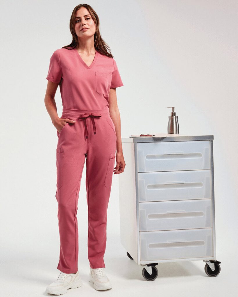 A woman is standing in the image, dressed in a pink top and pants. A white cabinet is situated behind her, and a bottle is placed on top of the cabinet.