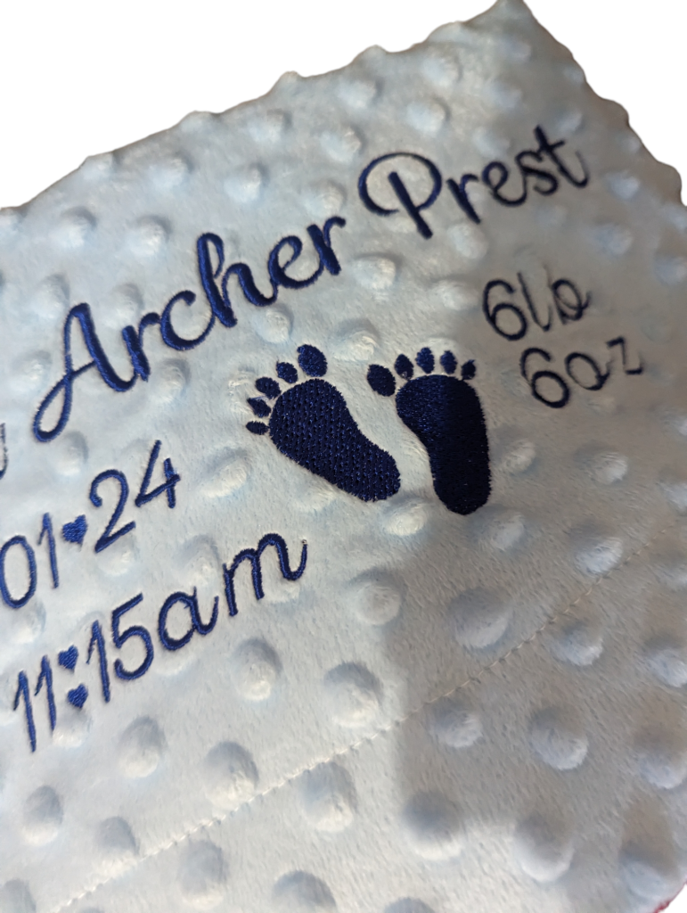 A white and blue blanket is depicted in this image, featuring a baby's foot and the name "archer" written in blue. The blanket is placed on a white surface.