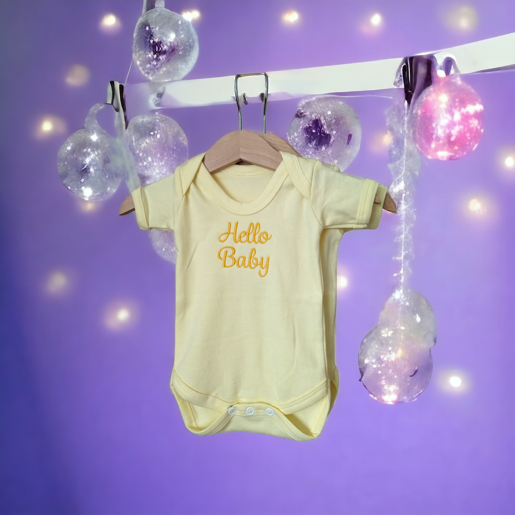 A yellow baby vest is depicted in this image, featuring the word "hello baby" written on it. The vest is hanging on a hanger, and a purple background is visible behind it.