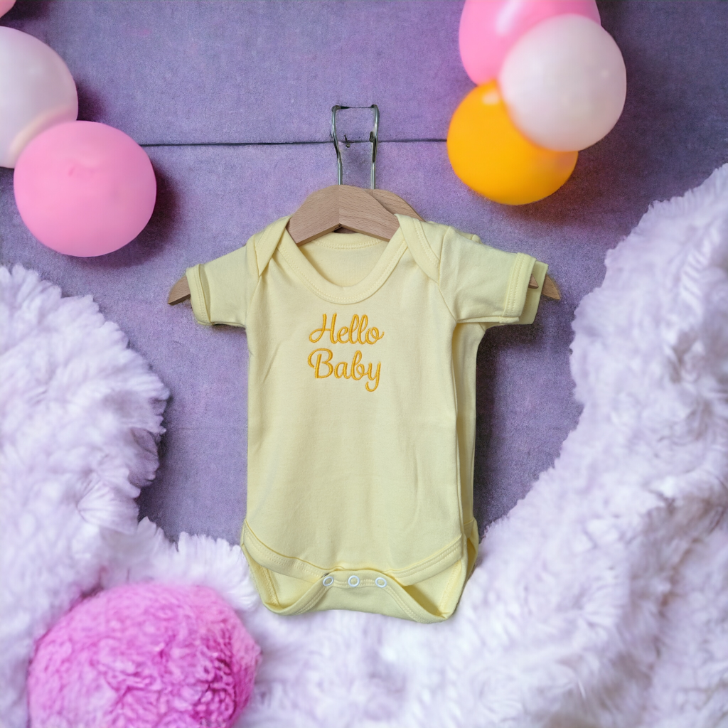 A yellow baby vest is depicted in this image, featuring the word "hello baby" written on it. The vest is hanging on a hanger, and a pink ball is situated to the left of the image.