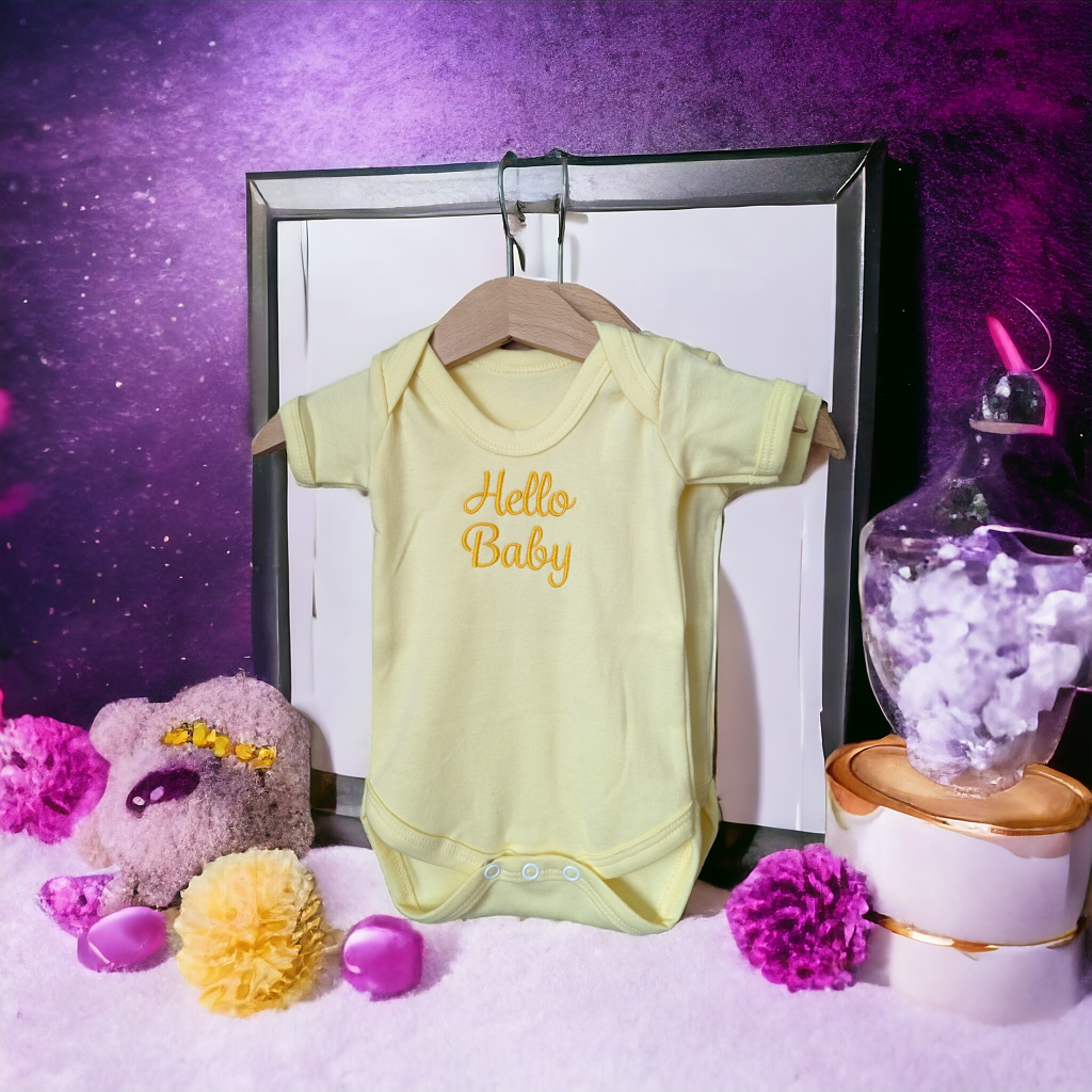 A yellow baby vest is displayed in the image, hanging on a hanger. A pink teddy bear is situated beside the vest. A white box containing flowers is placed on the floor to the right of the vest.