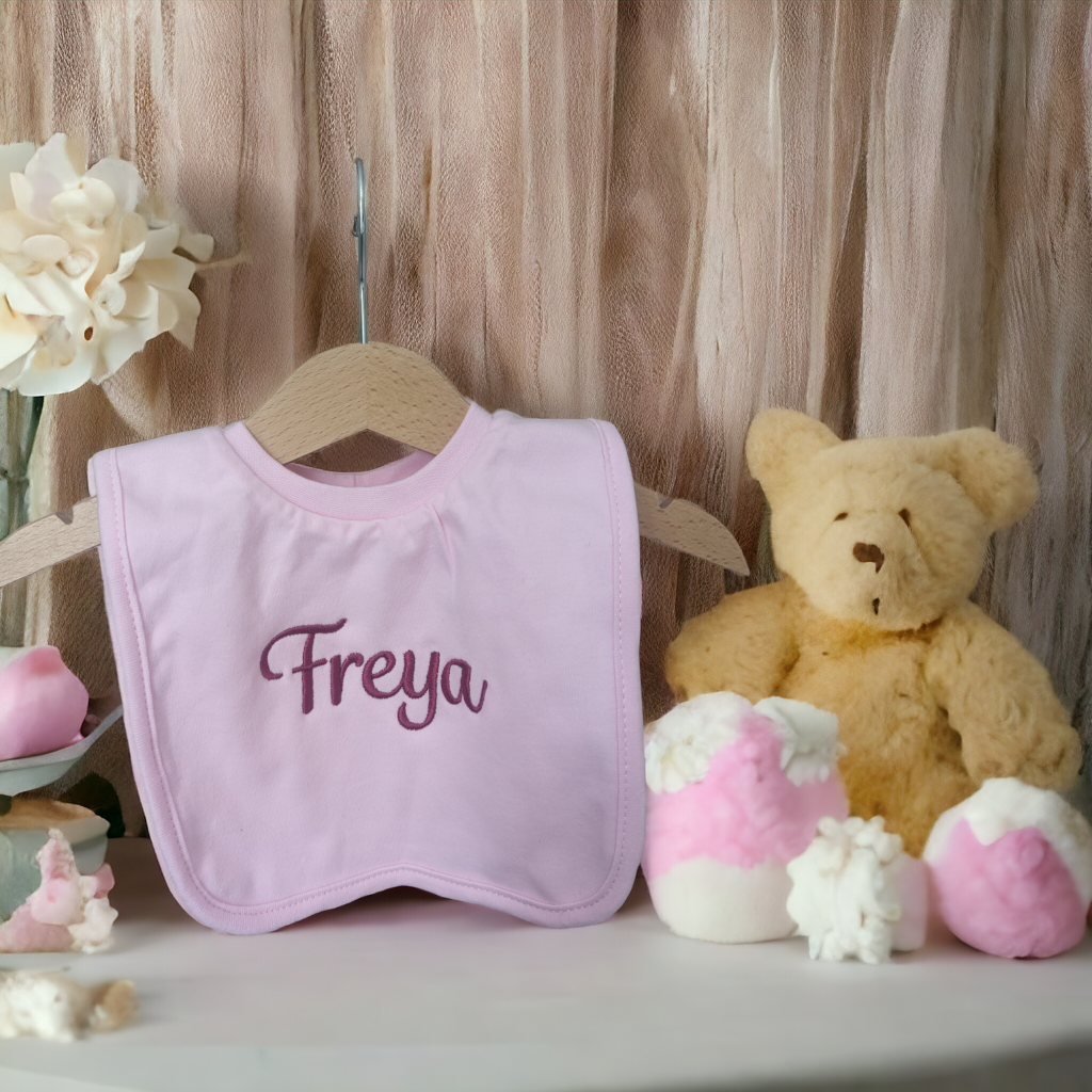 A pink bib is the main focus of this image, with a teddy bear positioned behind it. A pink curtain is visible in the background, and a white flower is situated to the left.