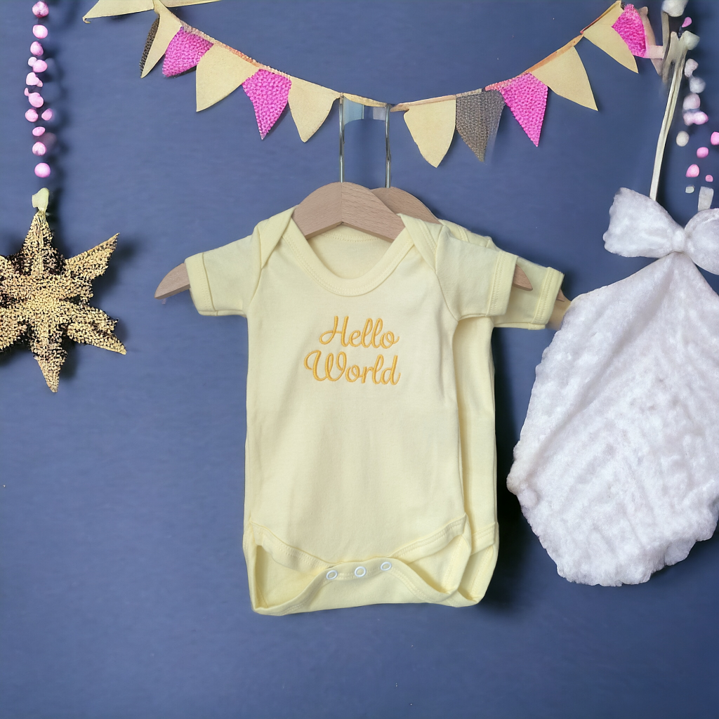 A yellow baby vest is depicted in this image, featuring the word "hello world" written on it. The vest is hanging on a hanger, and a white cloth is also present in the scene.