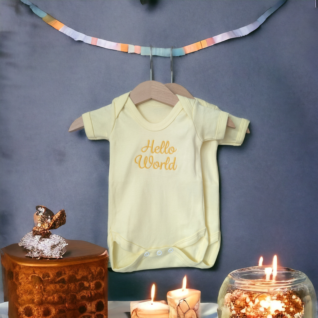 A yellow baby vest is displayed on a hanger, with a candle and a glass jar in the foreground. The background features a blue wall, and a ribbon is also present.