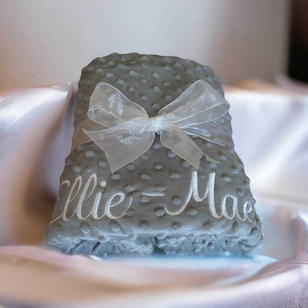 A grey-colored baby blanket is depicted in this image, with a white ribbon tied around it. The ribbon features the name "ellie mae" in black letters. The background is white.