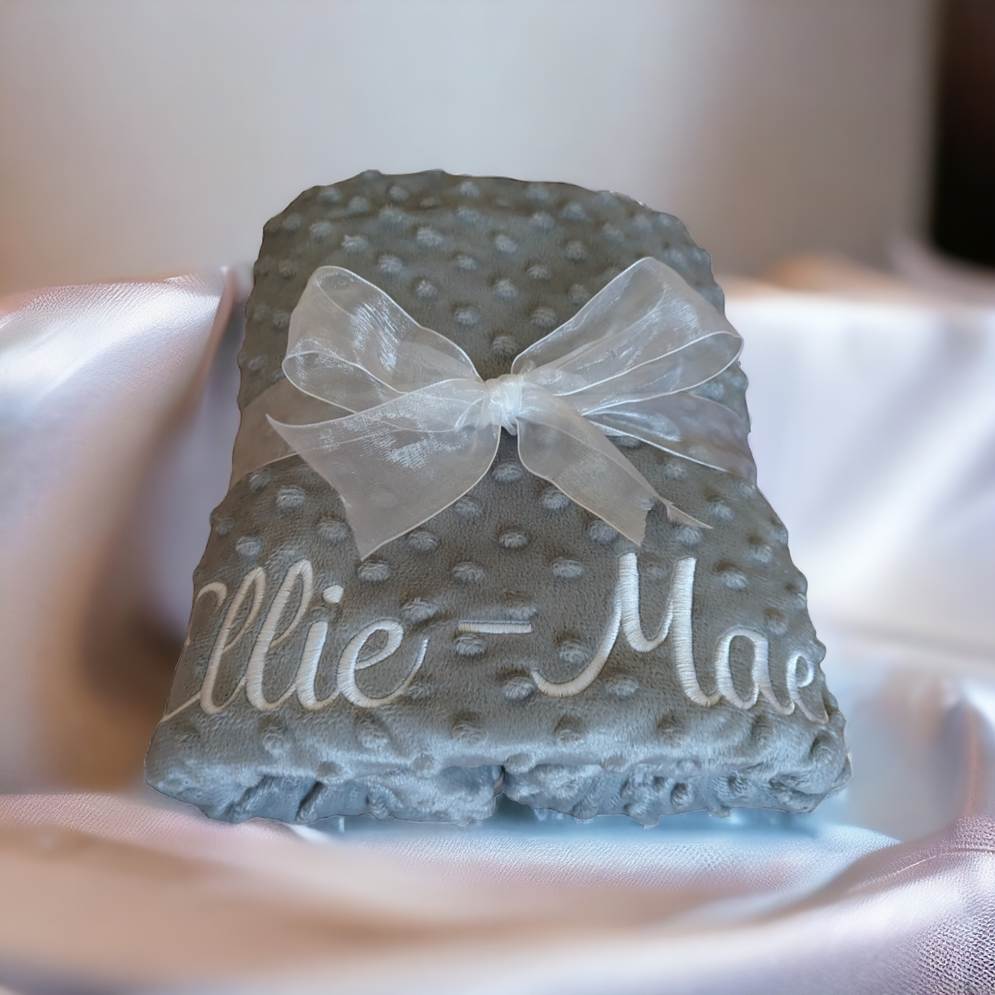 A grey-colored baby blanket is depicted in this image, with a white ribbon tied around it. The ribbon features the name "Ellie Mae" in black letters. The background is white.