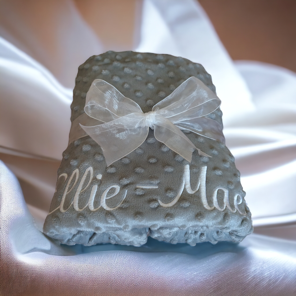 A grey-colored blanket is depicted in this image, with the name "ellie" written on it. The blanket is adorned with a white ribbon and a bow, and it is placed on a white cloth.