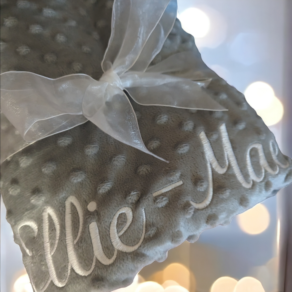 A grey-colored blanket is depicted in this image, with the name "ellie mae" embroidered on it. The blanket is adorned with a white ribbon and a bow.