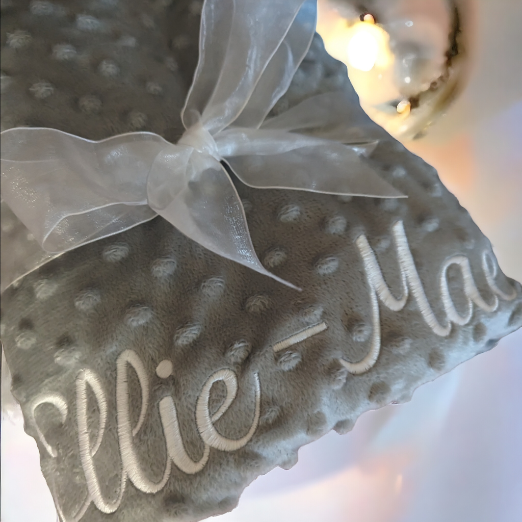 A grey-colored blanket is depicted in this image, with the name "ellie mae" embroidered on it. The blanket is adorned with a white ribbon and a bow.