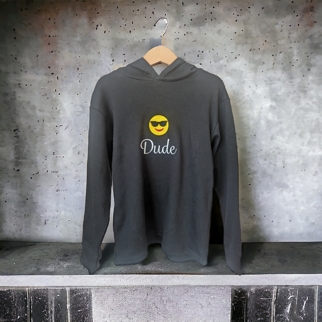 A hoody is displayed in this image, featuring a smiley face and the word "dude" written on it. The hoody is hanging on a hanger, and a wall is visible in the background.