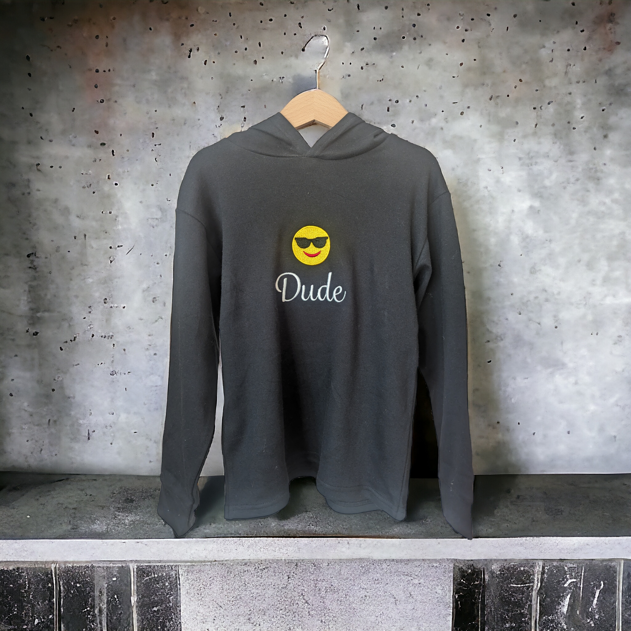 A hoody is displayed in this image, featuring a smiley face and the word "Dude" written on it. The hoody is hanging on a hanger, and a wall is visible in the background.