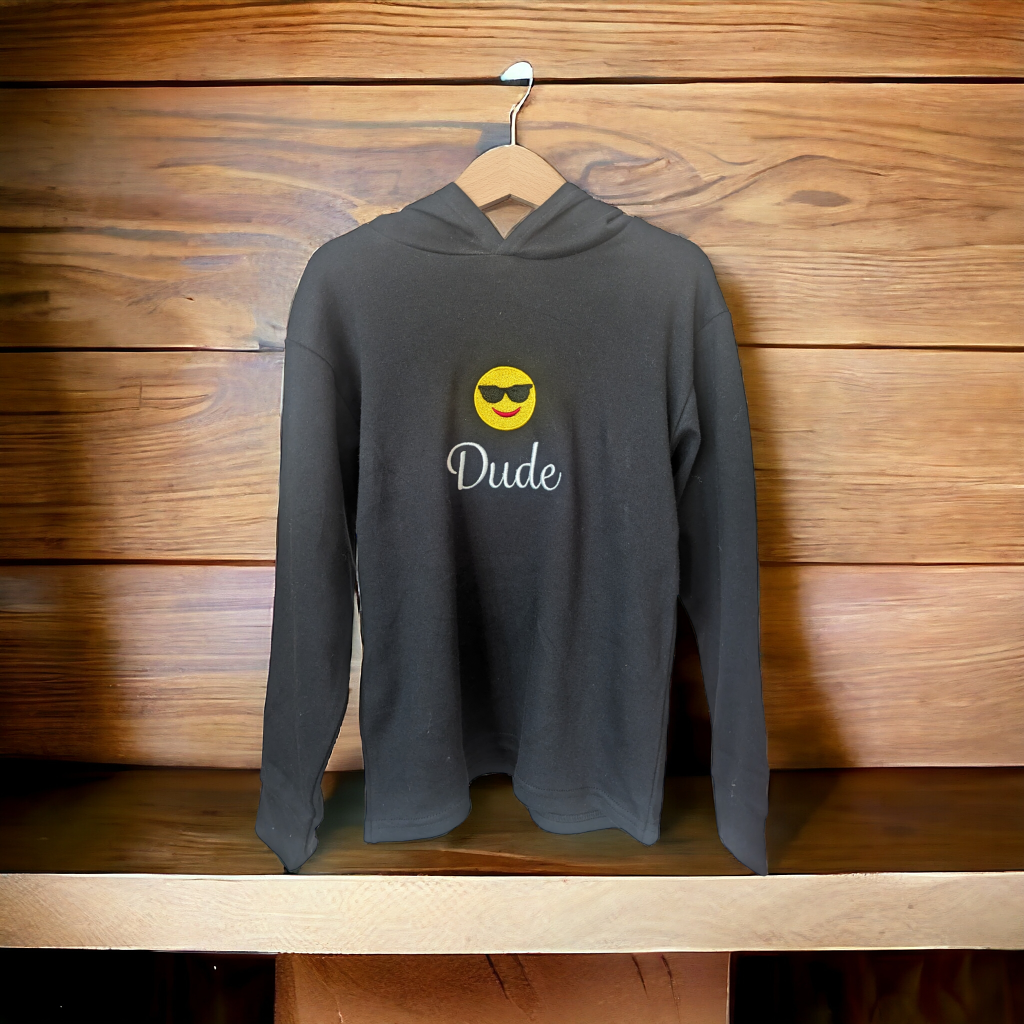 A black hoody is displayed in the image, featuring a white smiley face and the word "dude. " the hoody is hanging on a wooden surface.